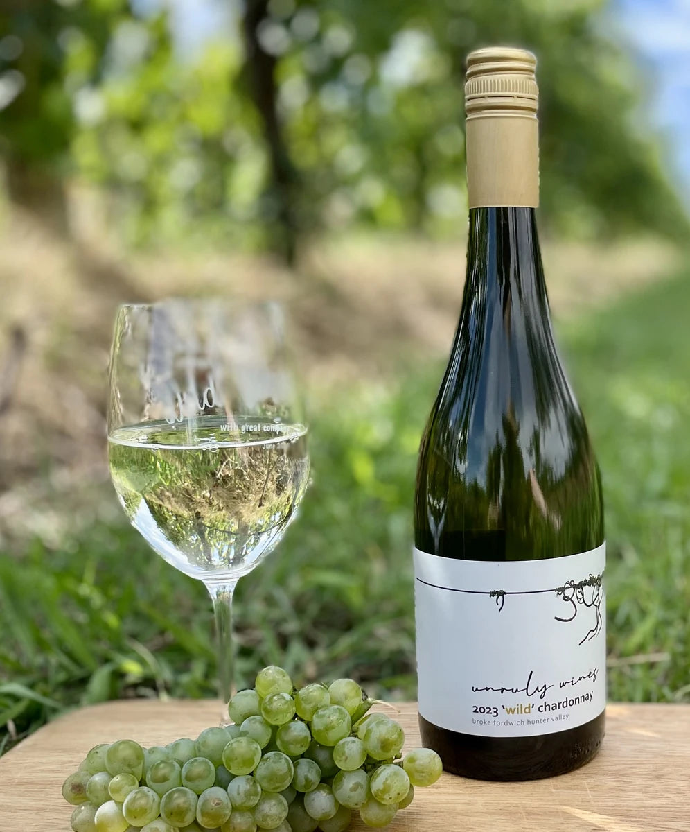 A glass and a bottle of Unruly Wines 2023 Wild Chardonnay on a barrel with chardonnay grapes, vines behind